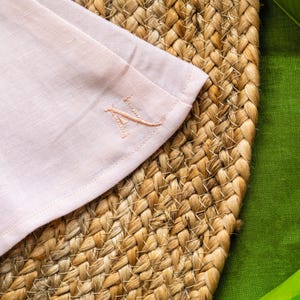 May include: A close-up of a pale pink linen napkin with an embroidered initial. The napkin is placed on a woven, circular placemat. A green fabric and a green leaf are in the background. The initial is in a matching pale pink thread.