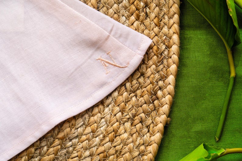 May include: A close-up of a pale pink linen napkin with an embroidered initial. The napkin is placed on a woven, circular placemat. A green fabric and a green leaf are in the background. The initial is in a matching pale pink thread.