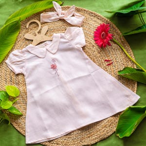 May include: A light pink baby dress with a flamingo embroidery, a matching headband, and a wooden bunny-shaped hanger. The dress has a Peter Pan collar and short sleeves. The items are arranged on a woven mat with green leaves and a pink flower.