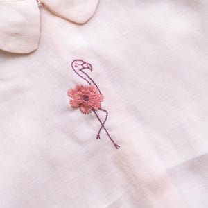 May include: Close-up of a light pink shirt with a Peter Pan collar. The shirt features an embroidered flamingo design with a pink flower. The embroidery is in shades of pink and purple.