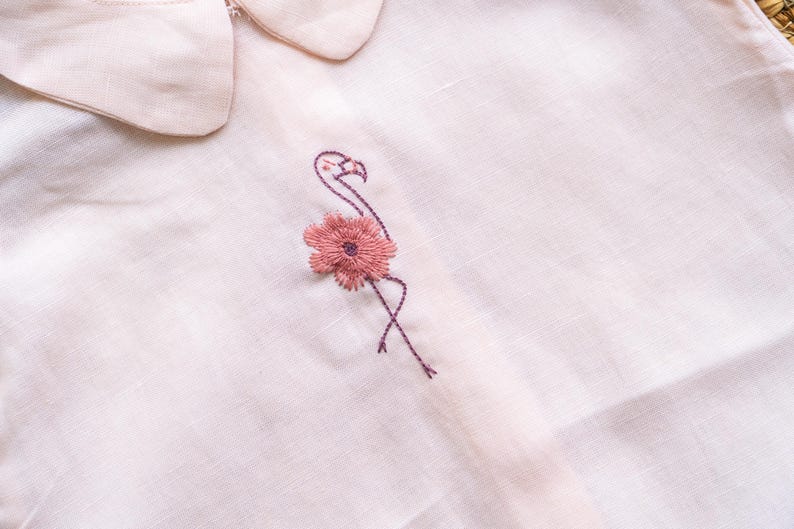 May include: Close-up of a light pink shirt with a Peter Pan collar. The shirt features an embroidered flamingo design with a pink flower. The embroidery is in shades of pink and purple.