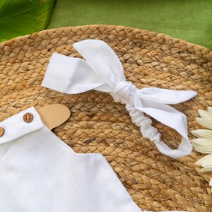 K&ouml;nnte beinhalten: Ein wei&szlig;es Baby-Outfit mit Holzkn&ouml;pfen und einem passenden Stirnband mit Schleife, das auf einer gewebten Matte liegt. Ein h&ouml;lzerner Hasenaufh&auml;nger und eine wei&szlig;e G&auml;nseblume sind ebenfalls zu sehen. Die Szene ist vor einem gr&uuml;nen Hintergrund.