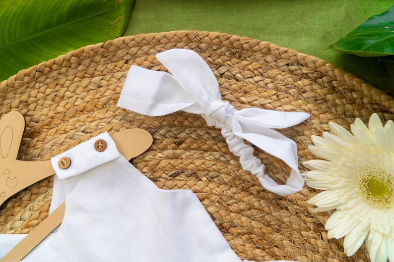 K&ouml;nnte beinhalten: Ein wei&szlig;es Baby-Outfit mit Holzkn&ouml;pfen und einem passenden Stirnband mit Schleife, das auf einer gewebten Matte liegt. Ein h&ouml;lzerner Hasenaufh&auml;nger und eine wei&szlig;e G&auml;nseblume sind ebenfalls zu sehen. Die Szene ist vor einem gr&uuml;nen Hintergrund.