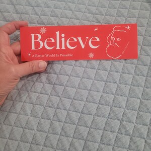 Puede incluir: Pegatina roja y blanca con la palabra "Believe" en texto blanco y la frase "A Better World Is Possible" en texto blanco m&aacute;s peque&ntilde;o. La pegatina tambi&eacute;n presenta una ilustraci&oacute;n estilizada de una persona con barba y copos de nieve.