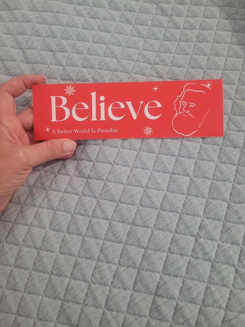 Puede incluir: Pegatina roja y blanca con la palabra "Believe" en texto blanco y la frase "A Better World Is Possible" en texto blanco m&aacute;s peque&ntilde;o. La pegatina tambi&eacute;n presenta una ilustraci&oacute;n estilizada de una persona con barba y copos de nieve.