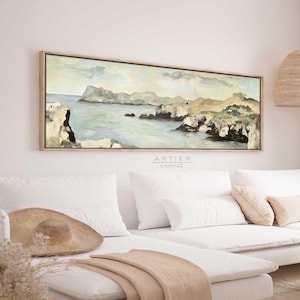 May include: A long, horizontal canvas painting depicting a seascape with rocky cliffs, green hills, and a blue ocean. The painting is framed in a light wood frame and hangs on a white wall above a white couch.