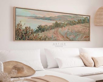 Puede incluir: Una pintura larga y horizontal de una escena de paisaje con una masa de agua en la distancia. La pintura está realizada en colores suaves y apagados, con tonos de rosa, azul y verde. La pintura está enmarcada en un marco de madera claro. La pintura está colgada en una pared blanca sobre un sofá blanco con almohadas y una manta.