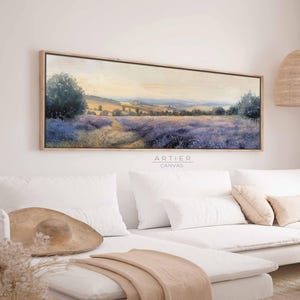 May include: A long, horizontal canvas painting depicting a landscape scene with a field of purple flowers, a winding path, and rolling hills in the distance. The painting is framed in a light wood frame and hangs on a white wall above a white sofa.
