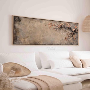 Pode incluir: Uma impressão em tela horizontal e longa com um fundo marrom opaco e um ramo delicado de flores rosa. A impressão é emoldurada em uma moldura de madeira clara e fica pendurada acima de um sofá branco com almofadas e uma manta.