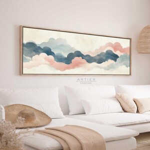 Op de afbeelding: Abstracte kunstprint met een aquarel schilderij van roze en blauwe wolken op een witte achtergrond. De print is ingelijst in een licht houten lijst en hangt aan een witte muur boven een witte bank.