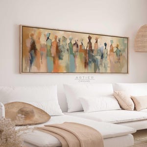 Puede incluir: Pintura abstracta de un grupo de personas en tonos apagados de marrón, azul, verde y naranja. La pintura está enmarcada en un marco de madera claro y cuelga de una pared blanca sobre un sofá blanco.