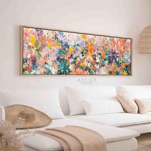 Puede incluir: Pintura abstracta con un diseño colorido y texturizado en tonos de rosa, naranja, amarillo, azul y verde. La pintura está enmarcada en un marco de madera claro y cuelga sobre un sofá blanco.