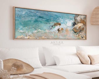 May include: A long, horizontal abstract painting of a seascape with blue and green water, white waves crashing on the shore, and brown rocks. The painting is framed in a light wood frame and is hanging on a white wall above a white sofa.