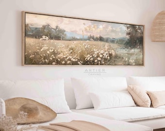 Puede incluir: Una pintura larga y horizontal sobre lienzo que representa un campo de flores silvestres blancas en un entorno paisajístico. La pintura tiene una paleta de colores suaves y apagados con tonos de verde, marrón y azul. La pintura está enmarcada en un marco de madera claro y está colgada en una pared blanca sobre un sofá blanco.