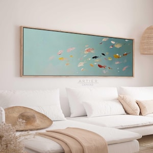 Könnte beinhalten: Ein großes abstraktes Gemälde von bunten Fischen, die in einem hellblauen Meer schwimmen. Das Gemälde ist in einem hellen Holzrahmen gerahmt und hängt an einer weißen Wand über einem weißen Sofa.