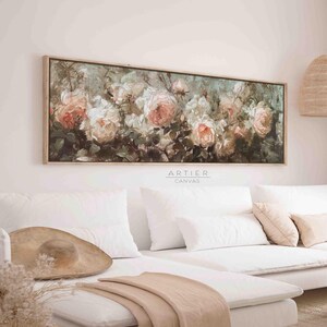 May include: A long, horizontal canvas painting of a floral arrangement featuring pink and white roses in a lush green setting. The painting is framed in a light wood frame and hangs above a white couch with pillows and a throw blanket.