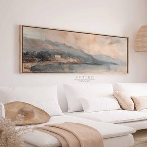 May include: A long, horizontal abstract painting of a landscape with mountains, water, and a sky with a soft pink and orange gradient. The painting is framed in a light wood frame and hangs on a white wall above a white sofa.