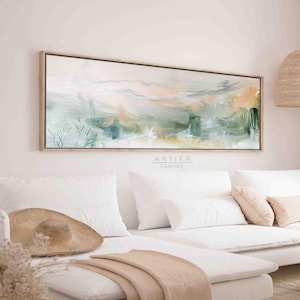 以下が含まれることがあります： 明るい木製の額縁に入った、長い横長の抽象画。絵画は、緑、ピンク、黄色の柔らかく落ち着いた色合いが特徴です。絵画は、枕と毛布のある白いソファの上に飾られています。