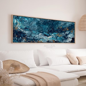 Pode incluir: Tela de arte abstrata com moldura de madeira. A obra apresenta uma técnica de impasto texturizada com tons de azul-petróleo, marinho, branco e dourado. A tela retangular está pendurada acima de um sofá branco com almofadas e um chapéu de palha. O texto "ARTIER CANVAS" é visível.