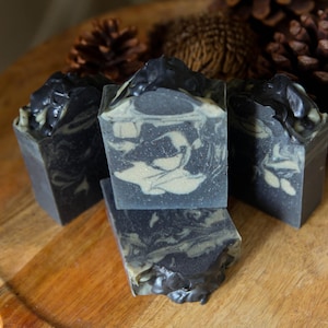 May include: Four square bars of handmade soap with a marbled design in shades of black, grey, and cream. The soaps are arranged on a wooden surface, with pine cones in the background.