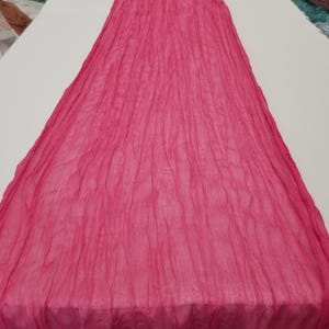 May include: A bright pink crinkled table runner made of a soft, sheer fabric. The runner is laid out flat on a white surface.