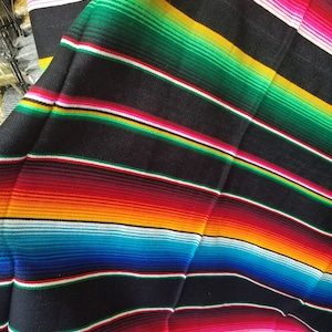 May include: A colorful Mexican blanket with stripes of red, pink, white, yellow, orange, blue, green, and black. The blanket is folded in half and has a fringe on the bottom.