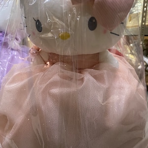 Puede incluir: Una gran muñeca de peluche blanca Hello Kitty con un vestido de tul rosa con purpurina. La muñeca tiene un lazo rosa en la cabeza y está envuelta en plástico transparente.