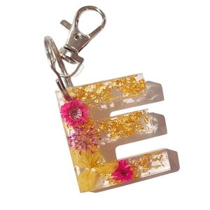 May include: A gold and white resin keychain with the letter "E" embedded with dried flowers and gold flakes. The keychain is attached to a silver split ring.
