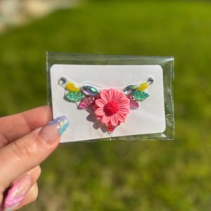 Pode incluir: Enfeite floral decorativo com um centro de flor rosa e detalhes coloridos. O design inclui pedras preciosas amarelas, verdes e iridescentes, apresentado em um cartão branco dentro de uma embalagem plástica transparente.