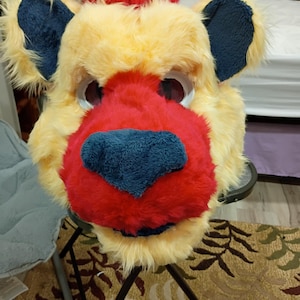 May include: A furry lion head costume with a red nose and a red tuft of fur on top. The ears are blue and the fur is yellow. The eyes are visible through the eye holes. This costume is suitable for cosplay or Halloween.