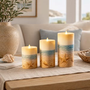 May include: A trio of LED pillar candles depicting a beach scene. The candles have a gradient design, with a sandy beach at the base, blue ocean in the middle, and a cream-coloured top. Starfish and shells are embossed on the base of each candle.