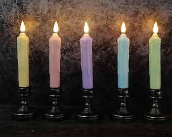 May include: Five multi-coloured LED taper candles in yellow, pink, purple, blue, and green, each set in a black metal candlestick. The candles have a realistic flickering flame effect, creating a warm ambiance. The background is a dark, textured surface.