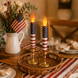 May include: Two battery-operated taper candles featuring a printed American flag pattern on the exterior. Each candle is topped with a glowing orange flame-shaped bulb and is supported by a polished gold-tone metal candlestick base.