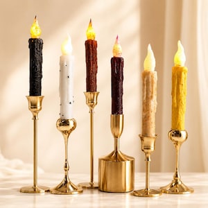 May include: Six LED taper candles in black, white, brown, dark purple, beige, and yellow, each featuring a textured wax-like finish and a glowing flame tip. The candles are held in individual gold-toned metal candle holders of varying heights.