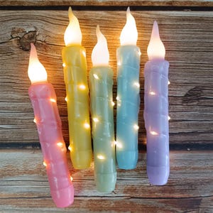 May include: Five pastel-colored LED candles with a spiral design. The candles are lit and have a warm glow. The candles are on a wooden surface.