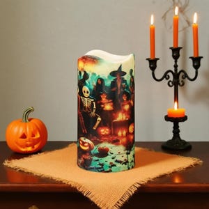 May include: A Halloween-themed candle with a spooky illustration of a skeleton, witches, and pumpkins. An orange carved pumpkin and a black candelabra with lit orange candles are also present. The candle sits on a burlap cloth.