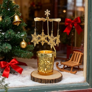 Könnte beinhalten: Ein goldfarbener Weihnachtskerzenhalter mit Schneeflocken-Design. Der Kerzenhalter steht auf einem Holzsockel und ist von einem Miniatur-Weihnachtsbaum, einer roten Schleife und einem kleinen Holzschlitten umgeben. Der Kerzenhalter hat eine rotierende Oberseite mit hängenden Schneeflocken-Ornamenten.