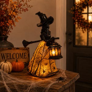 May include: A black witch figurine wearing a pointed hat and holding a broomstick. The dress features a glowing amber light with a silhouette pattern of bare tree branches and spiders. The witch holds a small, lit lantern in one hand.