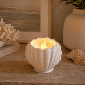 May include: A white seashell-shaped candle holder with a flickering LED light. The candle holder is made of a textured material and has a scalloped edge. The candle holder is on a wooden surface, with a painting and decorative items in the background.