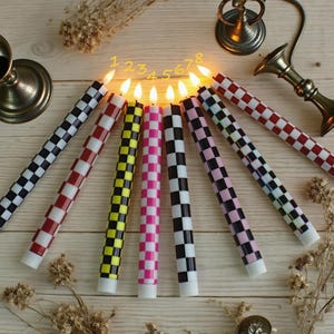 Puede incluir: Ocho velas encendidas con un patr&oacute;n de cuadros en varios colores, incluyendo negro, rojo, amarillo, rosa y blanco. Las velas est&aacute;n dispuestas sobre una superficie de madera clara. Tambi&eacute;n se ven candelabros decorativos de metal.