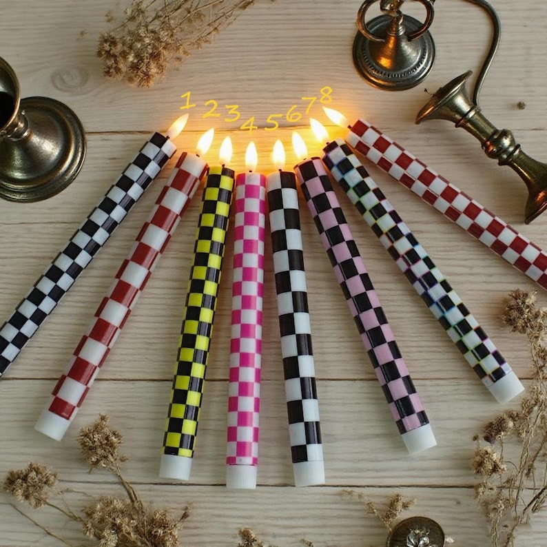 Puede incluir: Ocho velas encendidas con un patr&oacute;n de cuadros en varios colores, incluyendo negro, rojo, amarillo, rosa y blanco. Las velas est&aacute;n dispuestas sobre una superficie de madera clara. Tambi&eacute;n se ven candelabros decorativos de metal.
