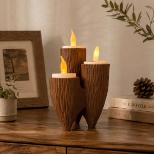 May include: A brown, textured candle holder shaped like three clustered tree trunks of varying heights. Each trunk features a flat top holding a white, cylindrical LED candle with a warm, glowing yellow flame-shaped light bulb on top.