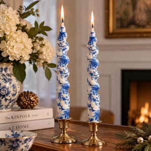 May include: Two tall, spiraled taper candles featuring a white base with a blue floral pattern. Each candle is lit, producing a small flame at the top, and is held upright by a polished brass candlestick holder with a circular base and stem.