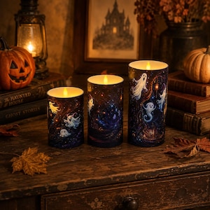 May include: Three cylindrical LED pillar candles featuring a dark, starry night sky print with various white cartoon ghosts. Each candle is topped with a warm, glowing faux flame, creating a soft light effect against the detailed, whimsical illustrations.