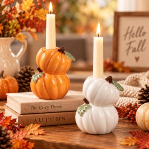 May include: Two ceramic candle holders shaped like stacked pumpkins, one orange and one white, each featuring a green leaf accent and a lit ivory taper candle. The holders are placed on two stacked books with the text The Cozy Home and AUTUMN.