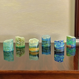 May include: A collection of six decorative LED candles, each with a unique painted design. The candles showcase artwork inspired by famous paintings, including landscapes and starry night scenes. Each candle is cylindrical and has a flickering LED light.
