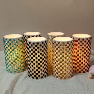 May include: Six cylindrical decorative candle holders with a mosaic design. The candle holders are in various colors, including teal, black, gold, silver, and red. Each candle holder has a white top and is illuminated from within.
