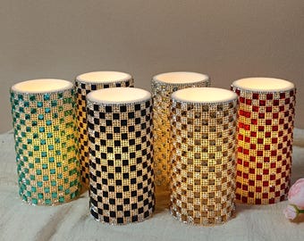 May include: Six decorative candle holders with a mosaic design. The cylindrical holders are in teal, black, gold, silver, and red. Each has a white top and is lit from within, creating a warm glow.