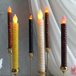 May include: Five decorative candles in different colors, including brown, yellow, white, black, and purple, are displayed on gold candle holders. The candles are twisted and have a flame-like tip.