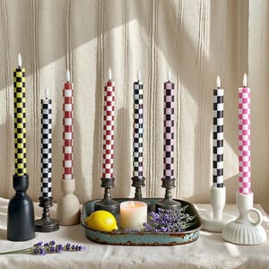 Puede incluir: Una colecci&oacute;n de velas c&oacute;nicas a cuadros en varios colores, incluyendo amarillo, negro, rojo, morado y rosa. Las velas se exhiben en diferentes candelabros, con un lim&oacute;n, una vela encendida y ramitas de lavanda en una bandeja.